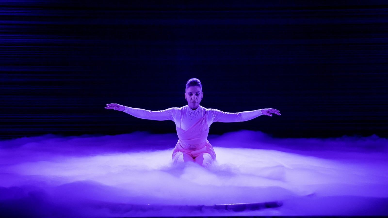 A dancer kneeling in a bed of low fog under purple light, 2023