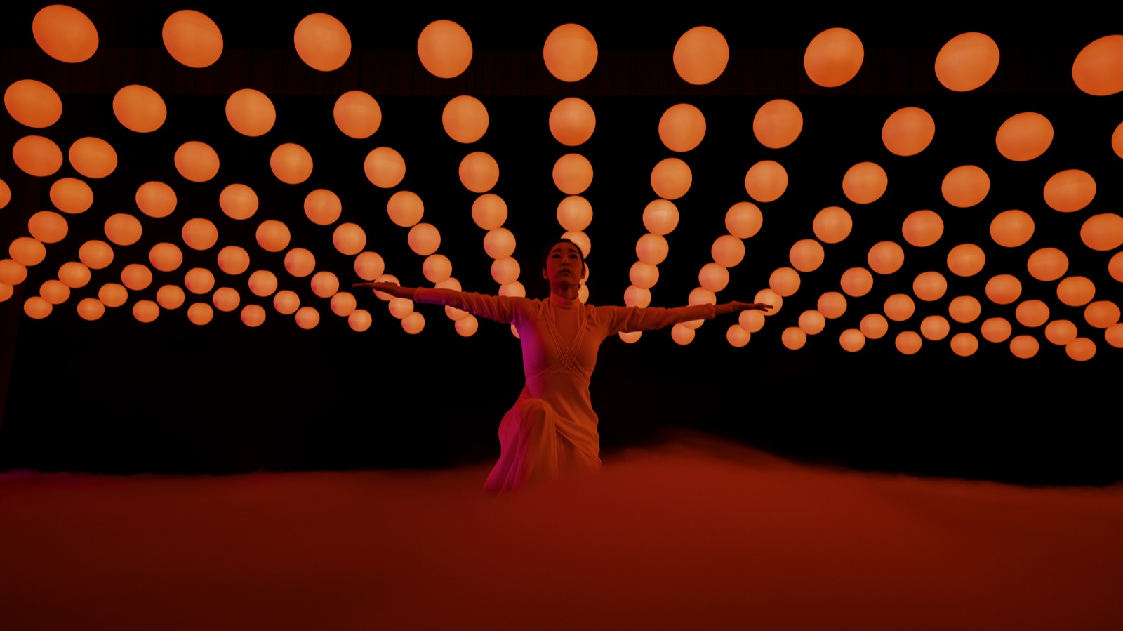 A dancer in low fog under a ceiling of orange light globes, 2023